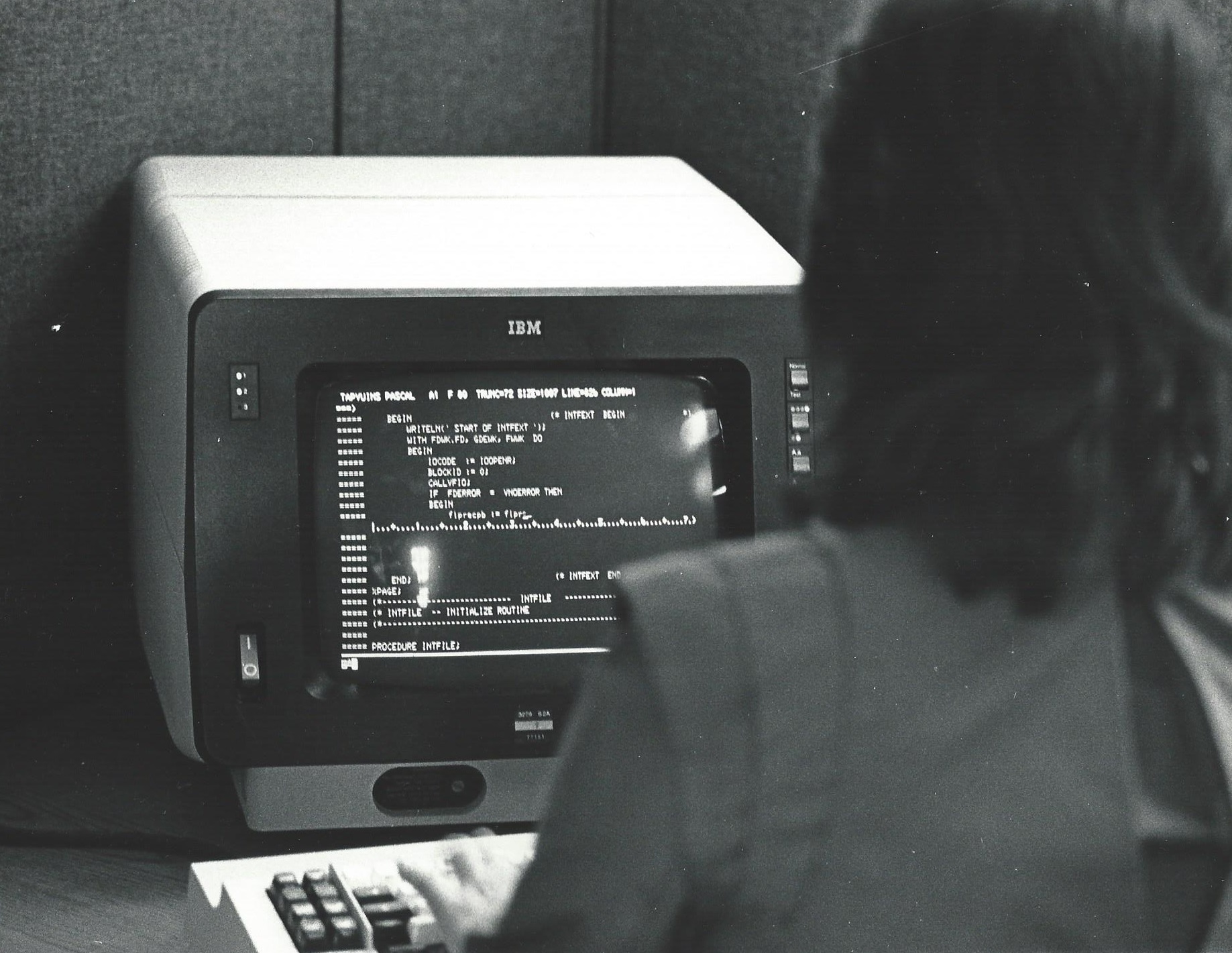 A programmer typing at an IBM terminal showing Pascal source code