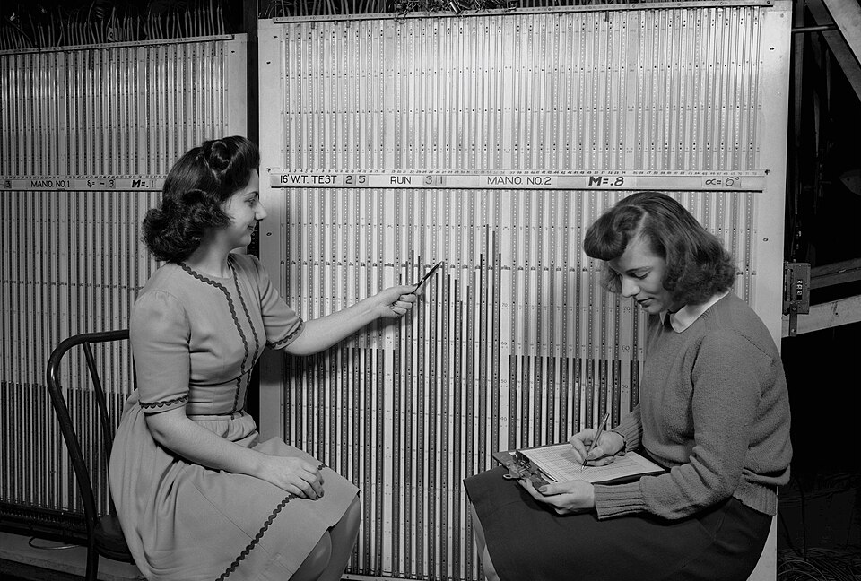 Two women reading a wind tunnel manometer board by hand